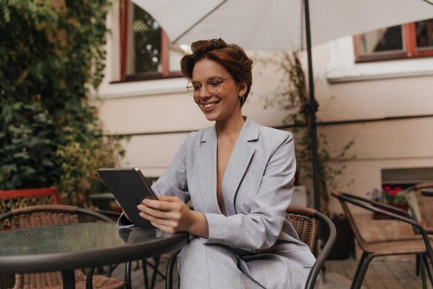 眼鏡の女性がストリートカフェに座ってタブレットでチャット灰色のスーツを着た幸せな女性がレストランで休憩し、ニュースを読む