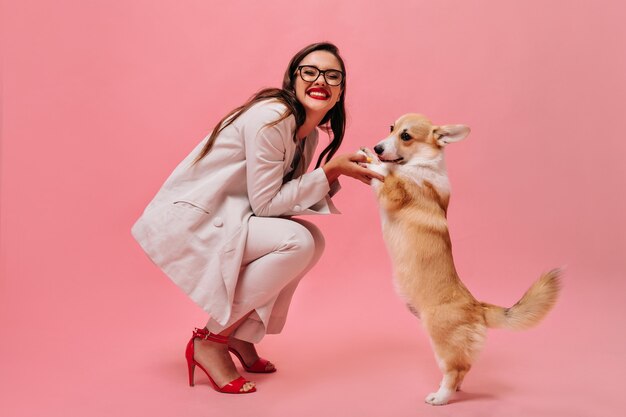 Lady in eyeglasses and suit plays with corgi on pink background.  Happy woman in office attire and red high heels smiles and holds corgi.