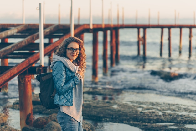 lady in gray hoodie and blue coat standing and smiling in beach during sunset