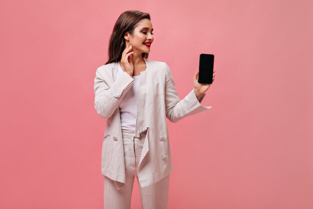 Lady in great mood holds smartphone on pink background.  Cute business woman in beige office suit looks at phone on isolated backdrop.