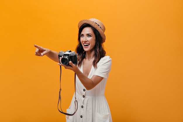 Lady in great mood is holding retro camera and pointing her hand against isolated background