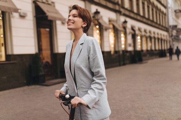 Lady in great mood riding scooter around city. Happy young woman in grey oversize jacket smiles and enjoys walk outside