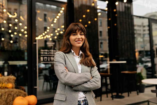 Lady in grey suit smiling at camera on background of stylish cafe with lights.