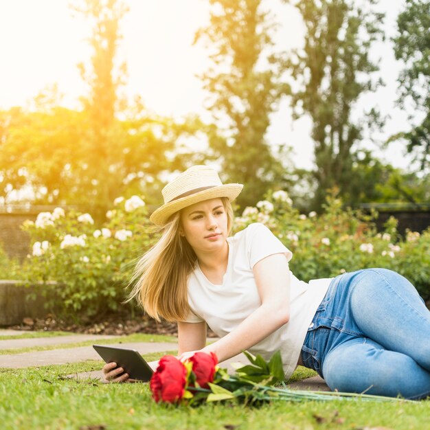 Lady in hat using tablet and lying on grass near flowers in park