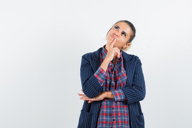 Lady holding finger on chin in shirt, jacket and looking pensive. front view.