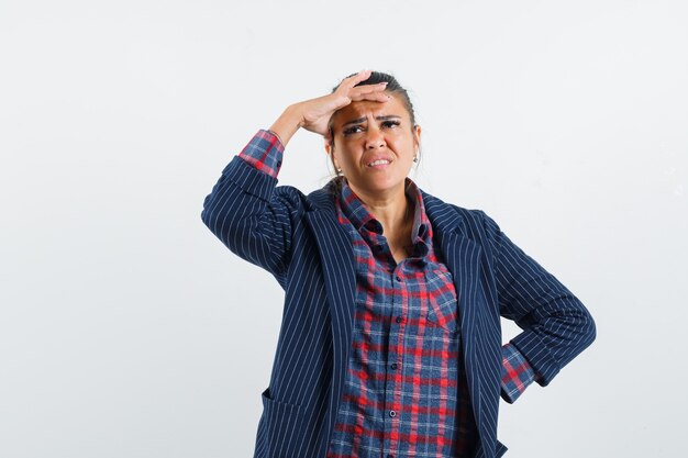 Lady holding hand on head in shirt, jacket and looking sorrowful , front view.