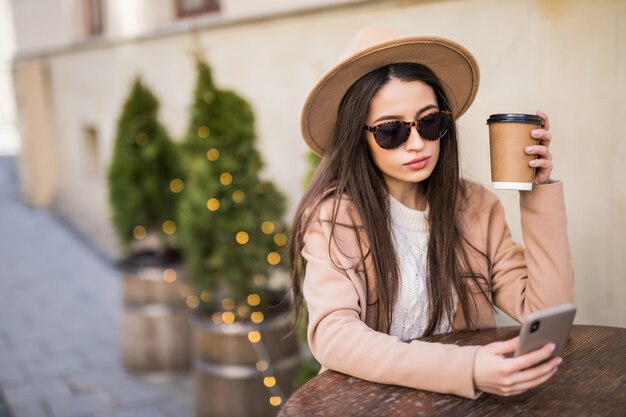 Lady is sitting on the table at cafe dresses in casual clothes dark sunglasses with coffee cup and phone