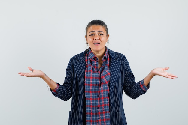 Lady making scales gesture in shirt, jacket and looking confused. front view.