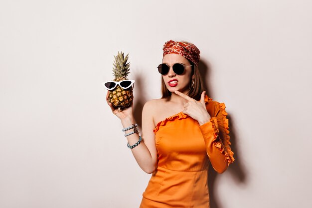 Lady in orange dress and sunglasses is posing thoughtfully and holding pineapple on isolated space.