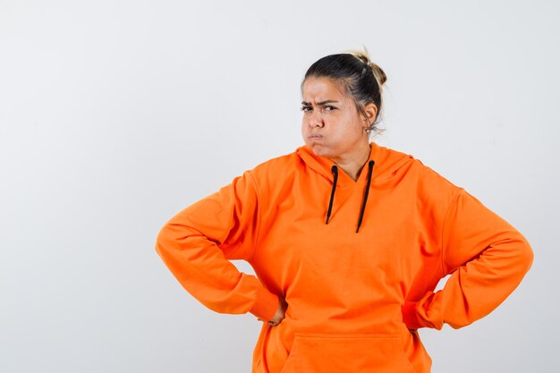 Lady in orange hoodie holding hands on waist and looking spiteful