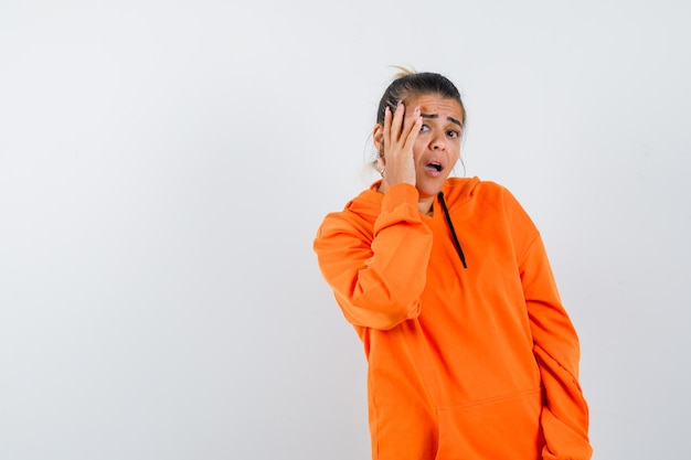 Lady in orange hoodie keeping hand on cheek and looking scared