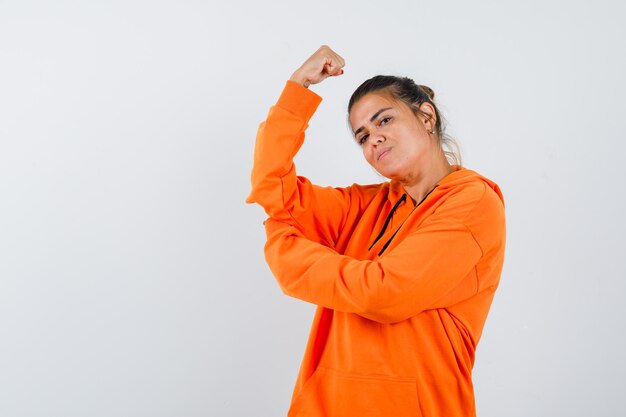 Дама в оранжевой толстовке с капюшоном показывает мышцы рук и выглядит сильной