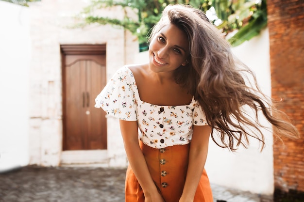 Lady in orange skirt and white top posing on cozy old European street Charming woman with long wavy dark hair is cute smiling