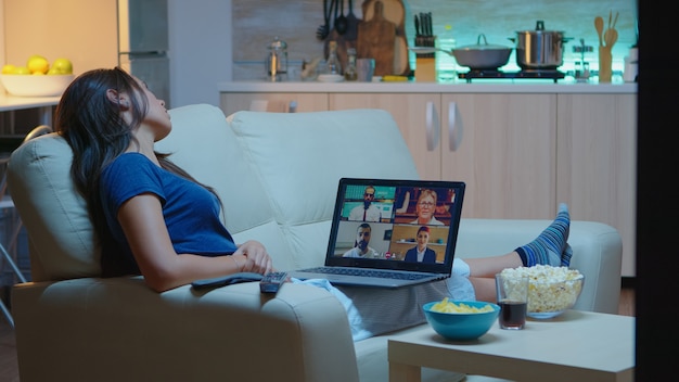 Lady in pajamas sitting on sofa having online meeting with project partners. Remote worker discussing at video conference consulting with colleagues using videocall and webcam working at laptop