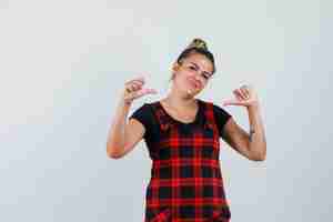 Free photo lady in pinafore dress pointing at herself with thumbs and looking proud , front view.