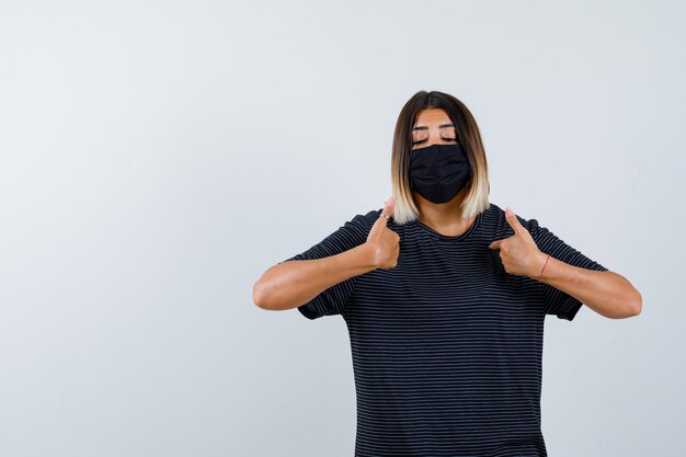 Lady pointing at herself in black dress, medical mask and looking proud. front view.