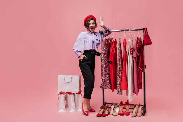 Lady poses in dressing room with bright clothes and shoes. Girl in beret and lilac blouse looking at camera on pink background.