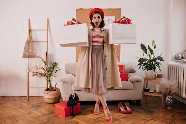 Lady in red beret, beige trench and dress holds shopping bags. Astonished young woman in long skirt and bright heels looks at camera.