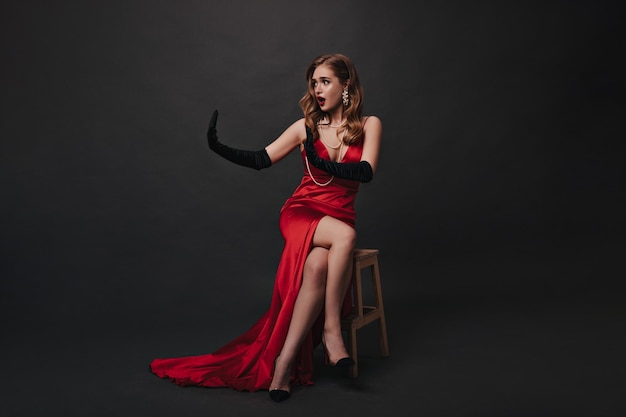 Lady in red long dress sitting on black background Superstar in luxury outfit posing on isolated backdrop