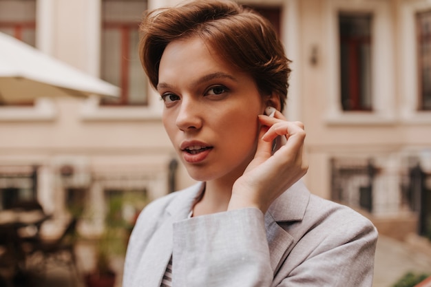 Free photo lady sensitively looks into camera and posing outside. close-up portrait of short-haired stylish pretty woman dressed in grey jacket. girl wearing wireless headphones