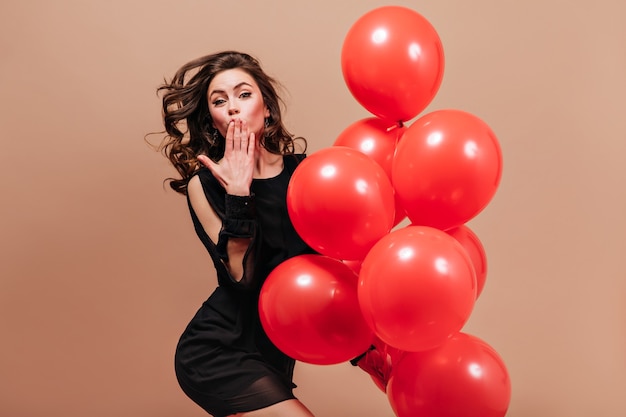 Lady in short black dress poses on beige background with balloons and blows kiss.