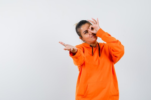 Lady showing ok sign on eye, stretching hand in orange hoodie and looking merry