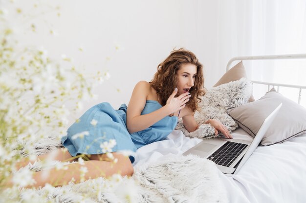 Lady sitting indoors on bed using laptop computer. Looking aside.