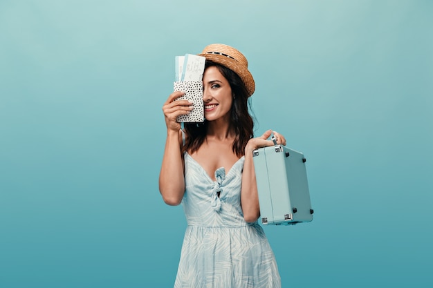 Lady in straw hat covers her face with tickets and holds suitcase against blue background. Dark-haired woman with short hair and big eyes posing.