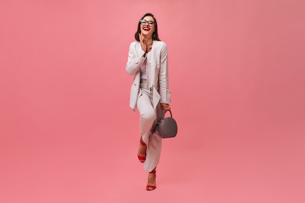 ピンクの背景に笑ってバッグを保持しているスーツの女性。眼鏡とカメラでポーズをとって赤い口紅で美しいビジネス女性。