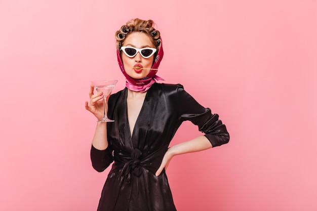 Lady in sunglasses posing on pink wall, holding martini glass and eating olive