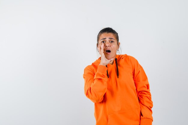 Lady telling secret with hand near mouth in orange hoodie and looking anxious