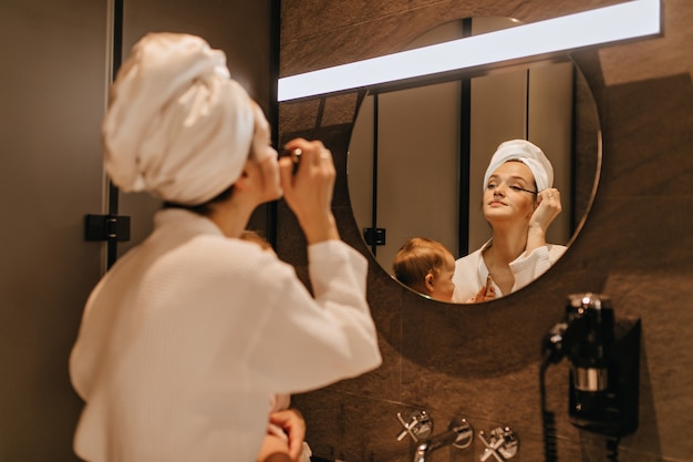 Lady in towel on her head does eye makeup, looking in bathroom mirror and holding baby.