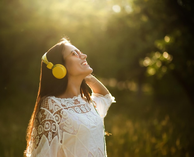 Леди в ярких желтых наушниках на природе. Веселая и счастливая молодая девушка стоит посреди лесной поляны и смотрит вверх в солнечных очках и ярко-желтых наушниках.