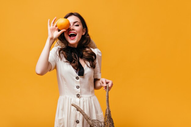 Lady in white dress laughs, covers her eye with orange and holds eco bag on orange background.