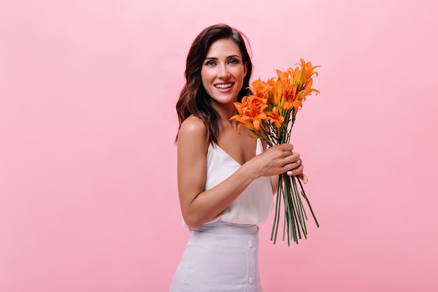 Lady in white outfit is smiling and holding bouquet of flowers. Beautiful woman posing for camera with cute orange flowers on isolated background.