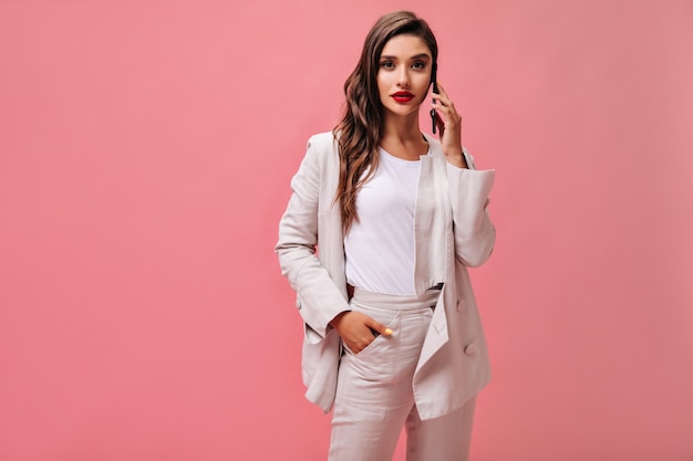 Lady in white suit looks into camera and talks on phone. Modern girl in light T-shirt and cream jacket poses on isolated background.