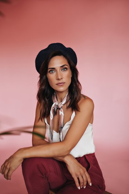 Lady in white top and silk scarf looks into camera on pink background. Green-eyed woman with short hair in red pants and light blouse looks at camera.