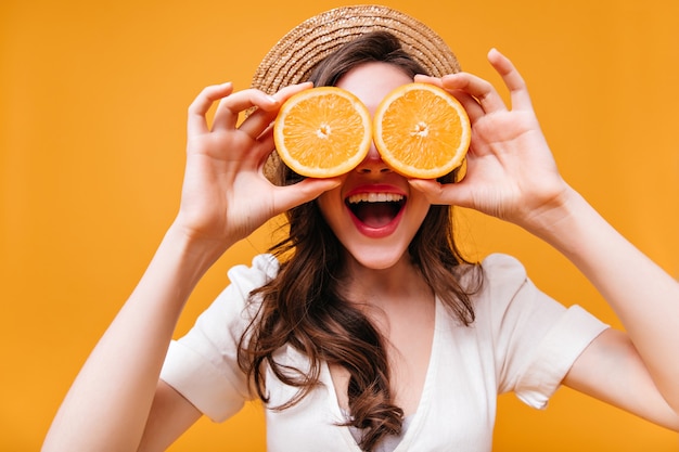 Lady in white top and straw hat laughs and covers her eyes with oranges.