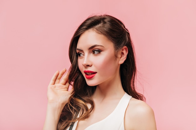 Lady in white top touches her wavy hair. Shot of brunette woman with red lips on pink background.