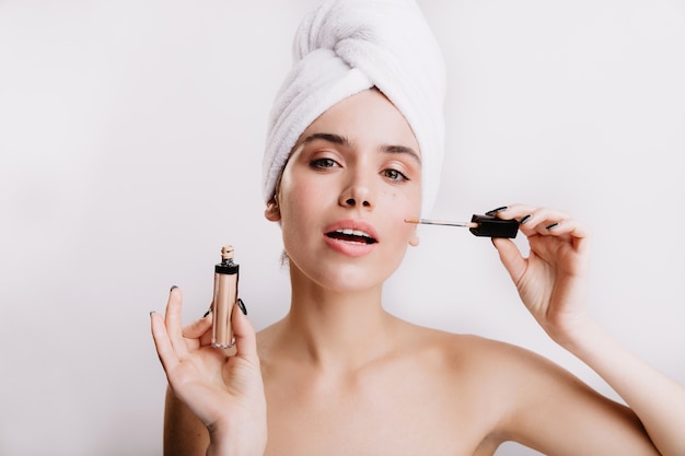 Lady in white towel on her head does nude makeup using concealer. Close-up portrait of green-eyed girl on isolated wall.