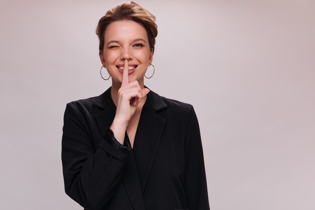 Lady winking and asking to keep secret on white backdrop. Happy short-haired woman in black jacket smiles on isolated background.