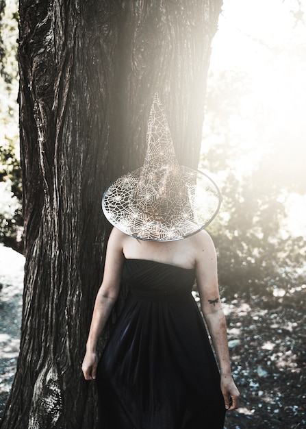 Free photo lady in witch costume with closed face by hat standing near tree