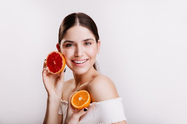 Lady with green eyes in white top posing on isolated wall without filters and makeup. Woman holds orange and grapefruit near face.