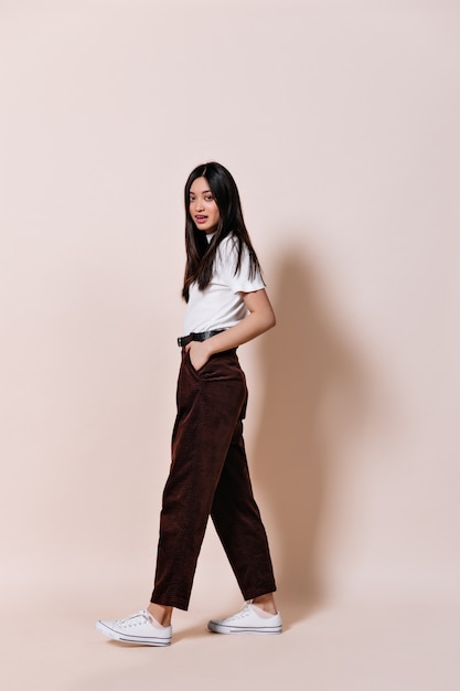 Lady with long hair dressed in brown pants posing on beige wall