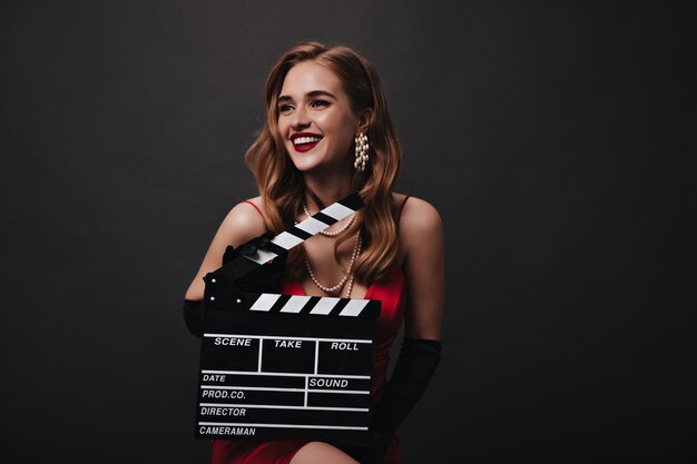 Lady with red lips holding clapperboard on black background Happy woman in luxury silk dress smiling and posing on isolated backdrop