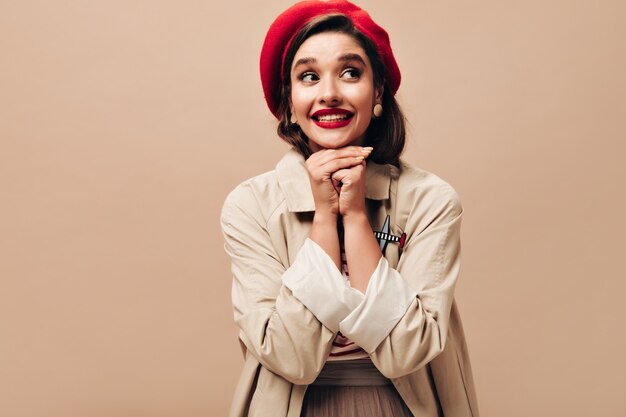 Lady with red lips smiling on beige background.  Funny young woman in bright beret, in cute earrings posing at camera on isolated backdrop.