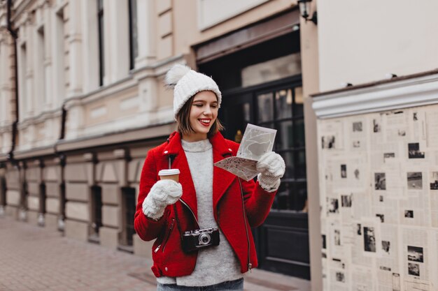 Lady with red lipstick dressed in white hat, gloves and short wool coat is holding glass of tea and paper card, posing outdoors with retro camera.