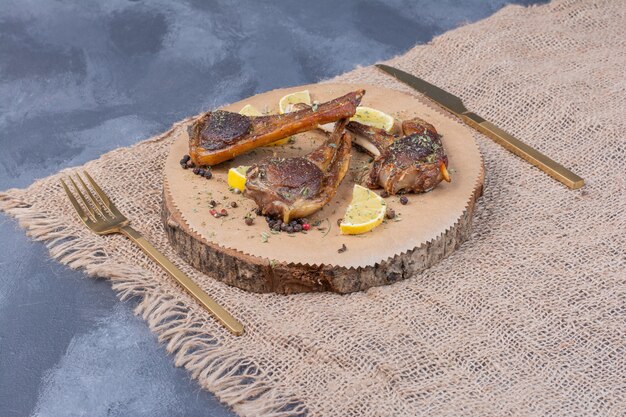 Lamb chomps on wooden board with lemon slices and cutlery on tablecloth.