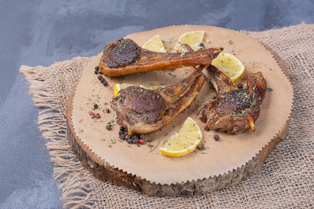 Lamb chomps on wooden board with lemon slices and cutlery on tablecloth.