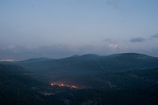 Landscape of hills covered in forests and lights under a cloudy sky during the evening